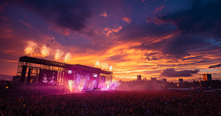 Crowd of people at outdoor festival concert with fireworks and vibrant sunset sky.